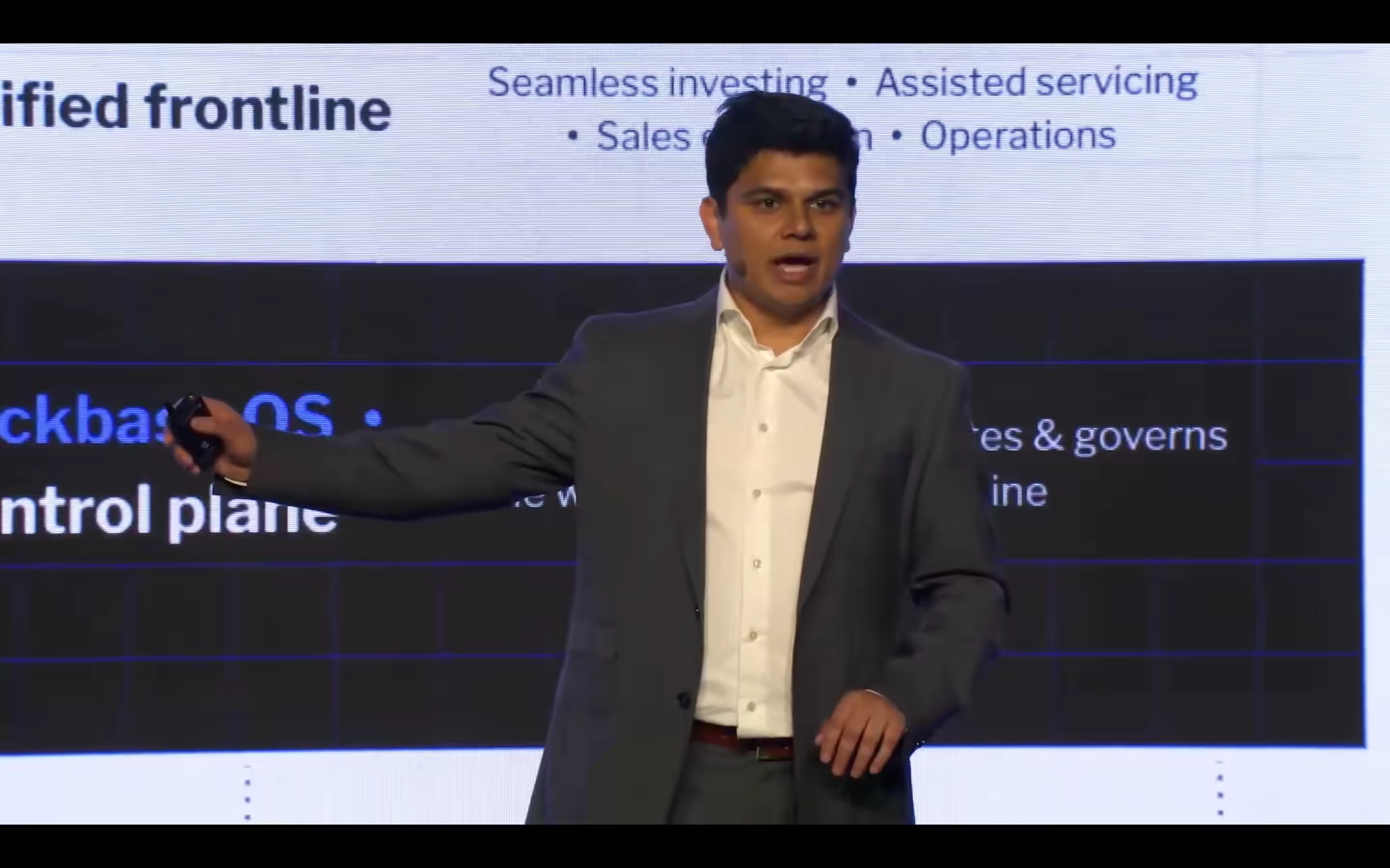 A speaker presenting on stage at a Backbase Engage event, gesturing toward a slide showing the Unified Frontline and Backbase OS architecture. Visible slide text includes use cases: Seamless investing, Assisted servicing, Sales optimization, and Operations, alongside references to the Banking OS control plane.