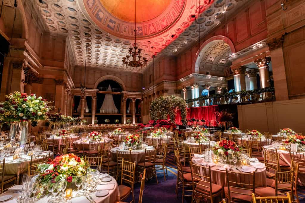 Elegant banquet hall set for a formal event with high domed ceilings, gold chiavari chairs, round tables with lush floral centerpieces, fine dining ware, and warm ambient lighting. Features a stage with red curtain and balcony decorated with greenery, perfect for corporate holiday parties.