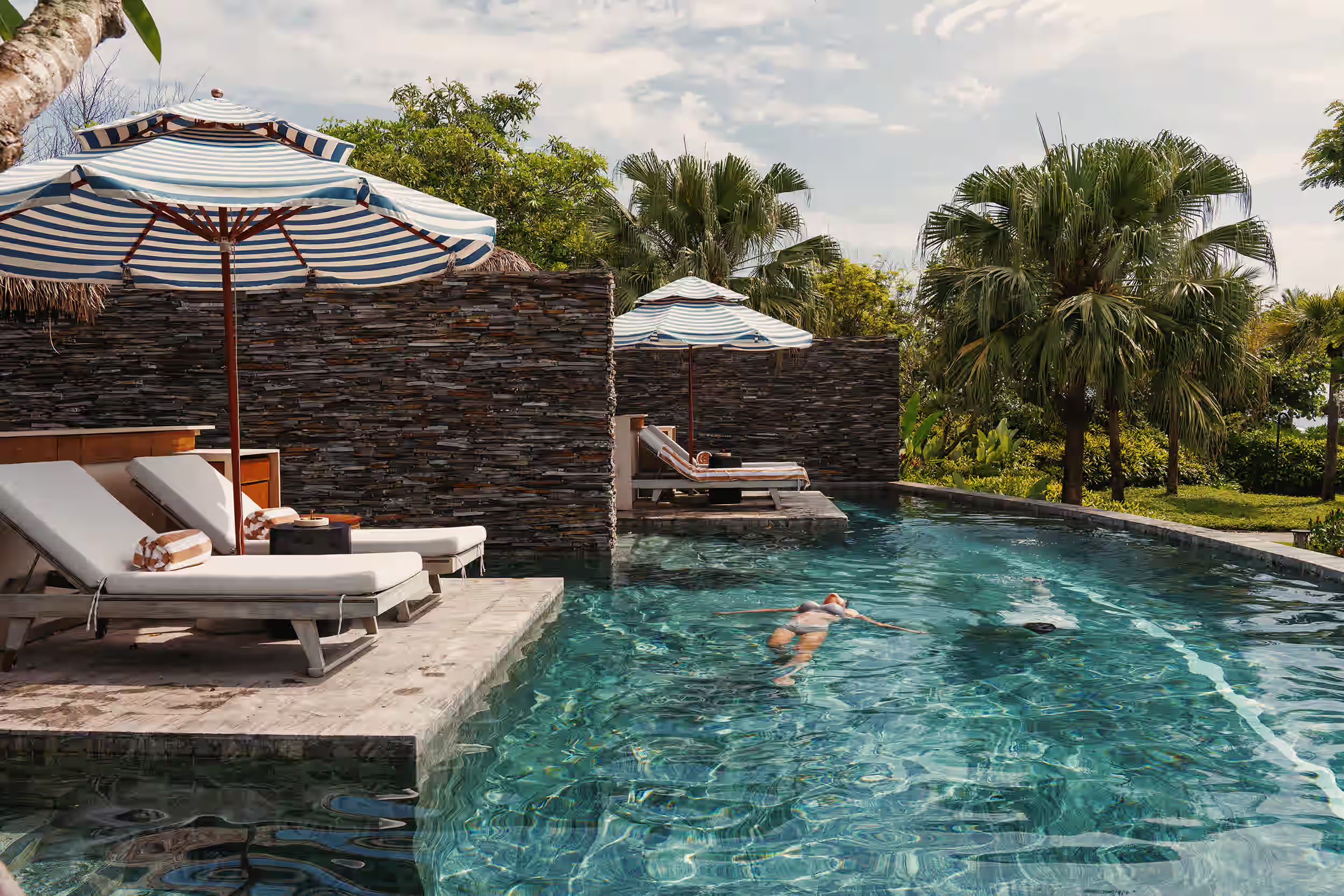A guest swimming beside shaded loungers and a private Hide.Out cabana surrounded by greenery.