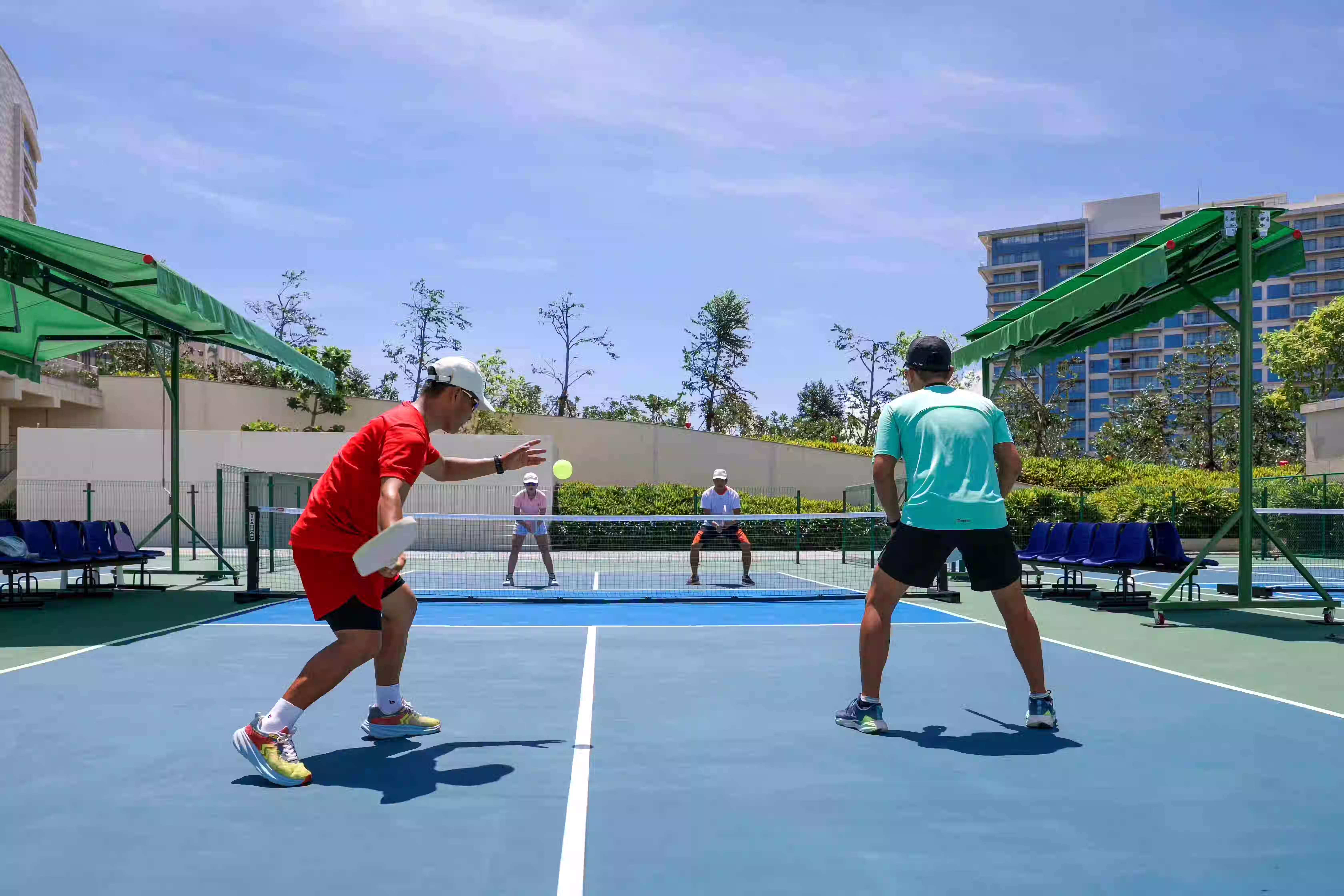 Pickleball players rallying on a sunlit Hoiana court framed by palm trees and resort buildings.