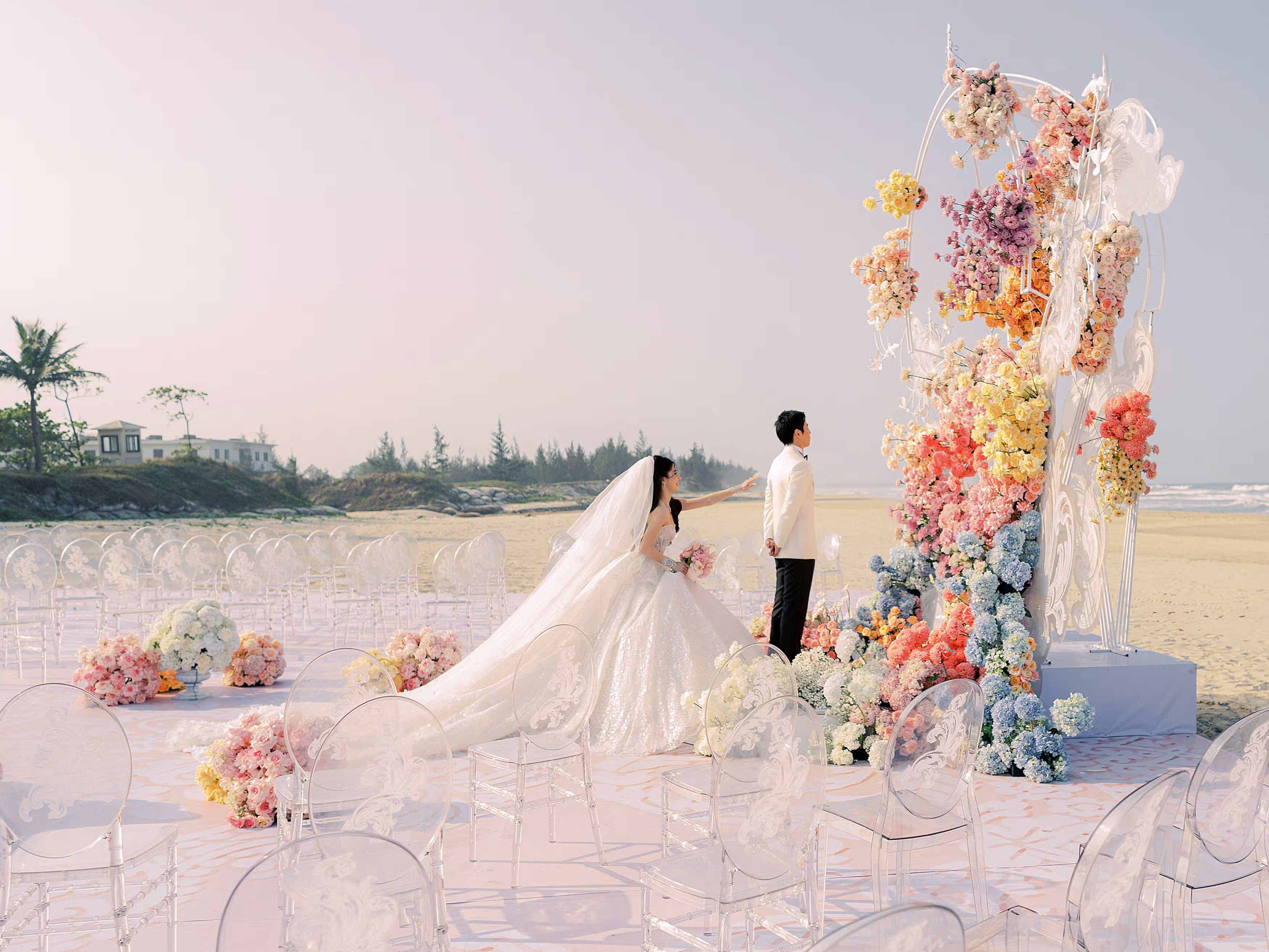 A newlywed couple stands beside sculptural peach-toned florals and pampas grass on the beach