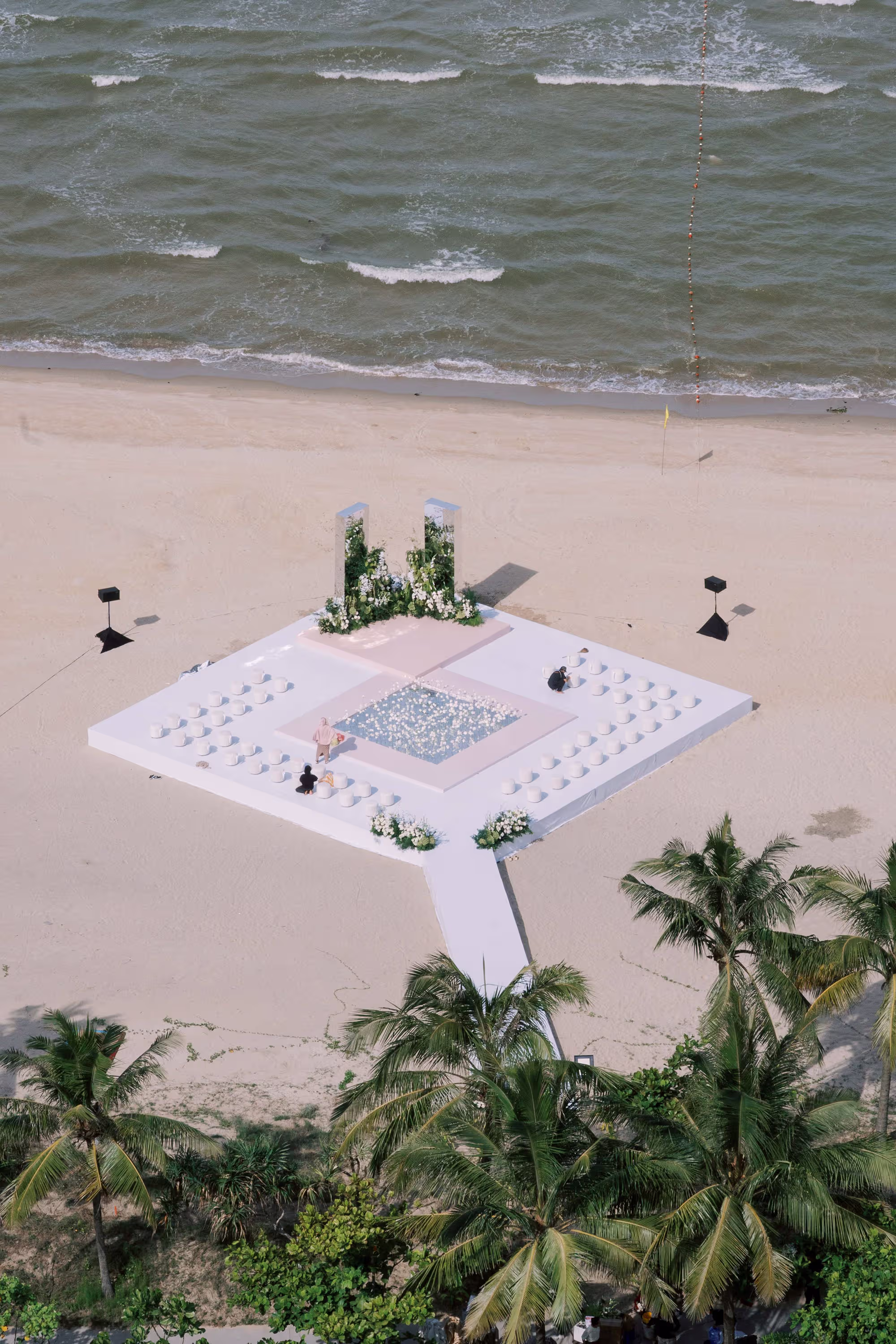 A minimalist white ceremony platform rests on the sand, framed by tropical palms and the ocean beyond
