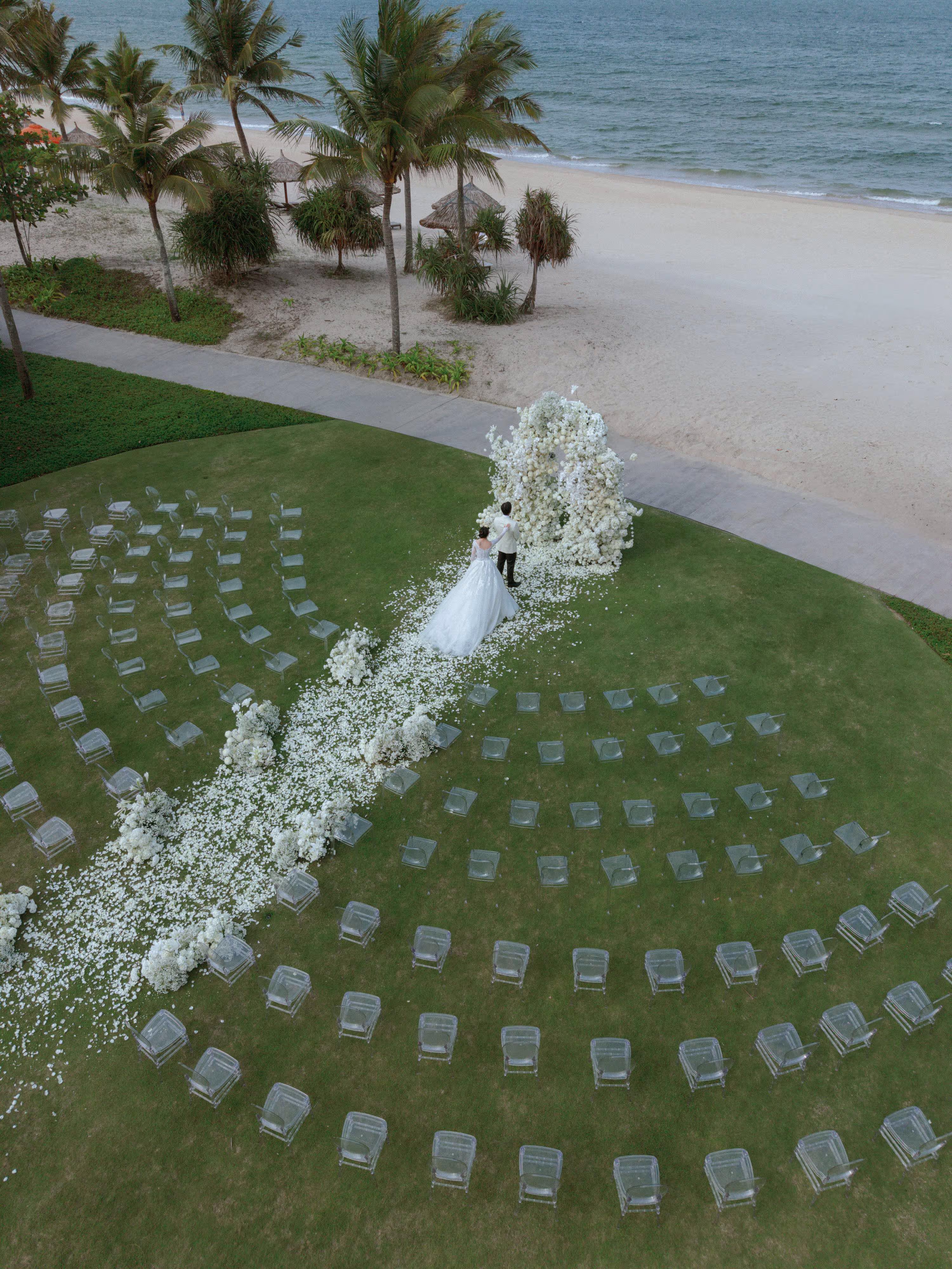 Oceanfront wedding ceremony at Beach Lawn, Hoiana, where a petal-lined aisle and curved rows of transparent chairs lead to a sculptural white floral backdrop beneath tropical palms.