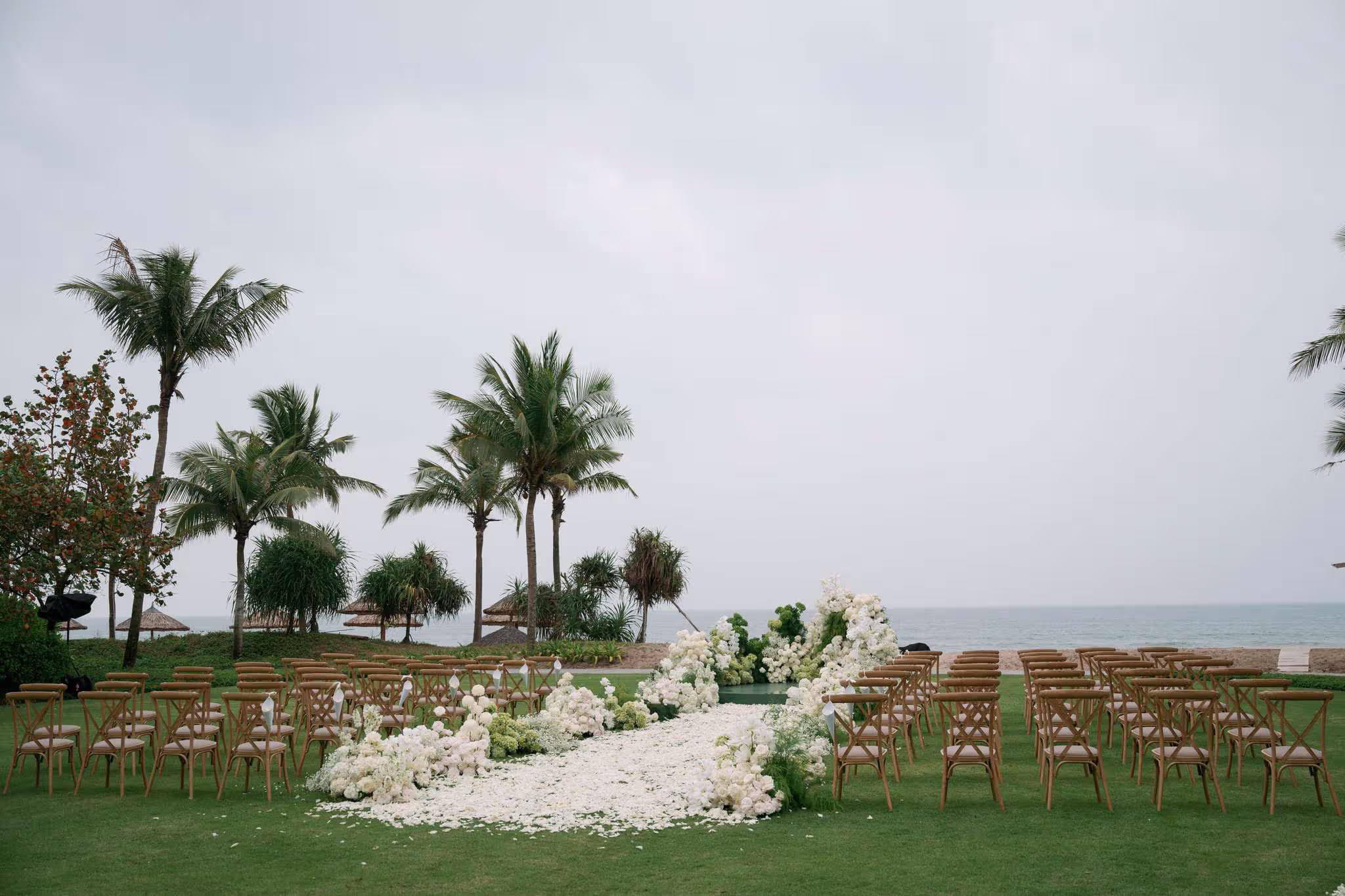 Wooden chairs and flowing white florals create an elegant wedding setting between tropical palms and the sea