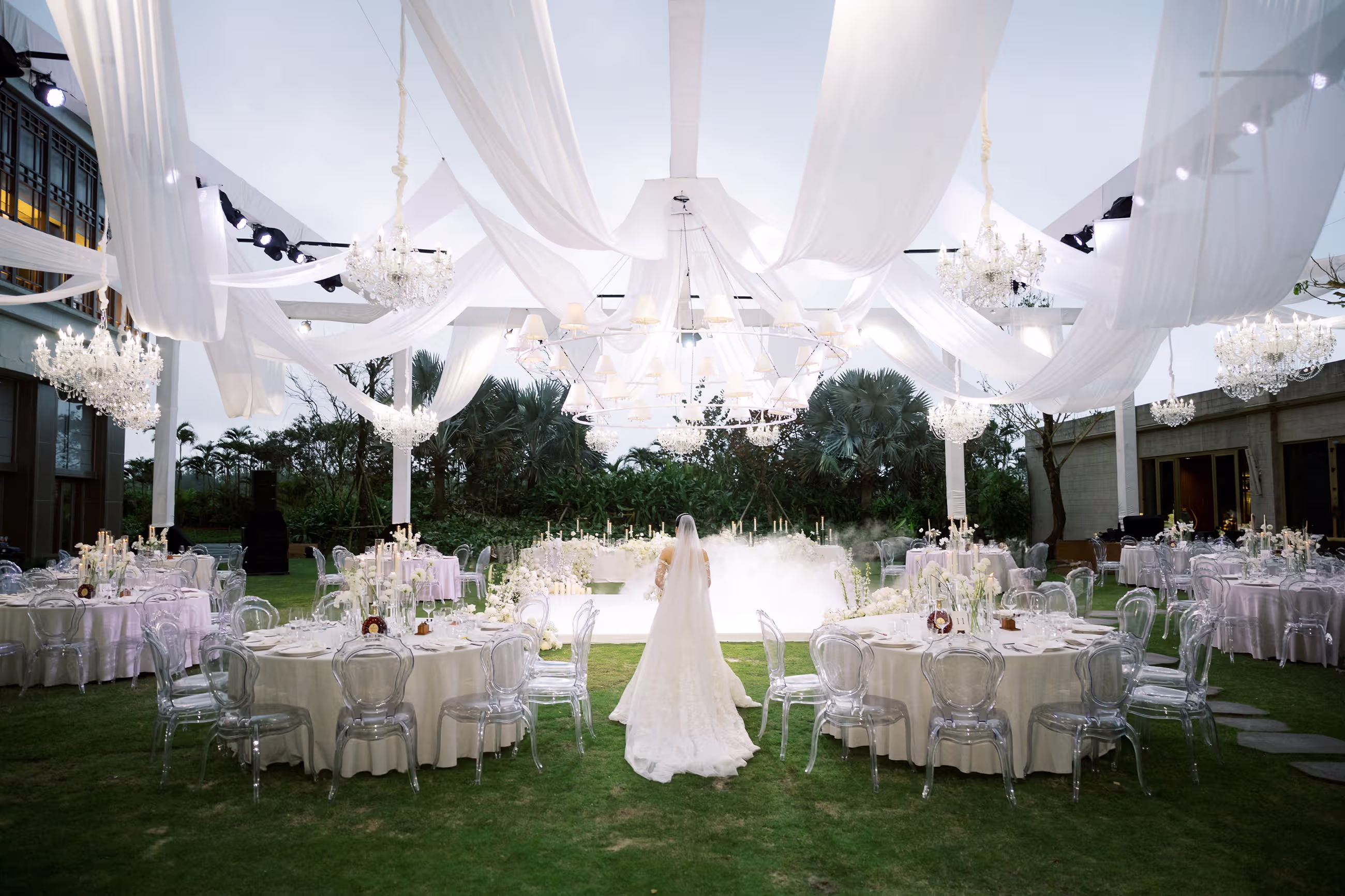 An elegant outdoor reception unfolds beneath billowing white drapes, with round banquet tables arranged across the garden lawn