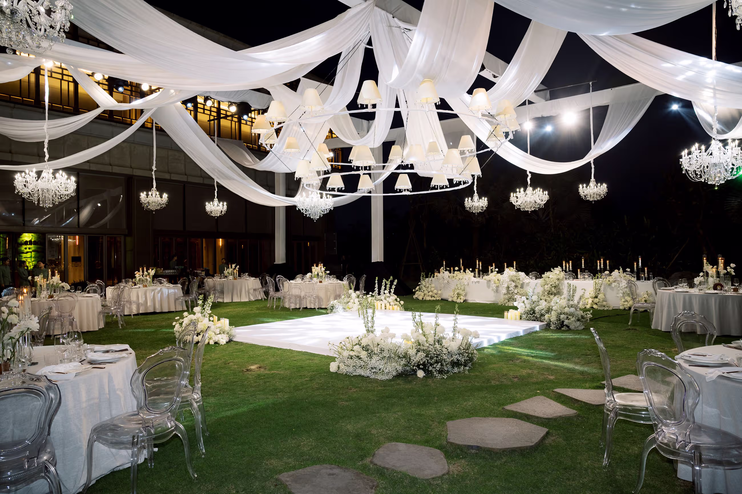 Soft lighting and flowing white fabric create an intimate evening reception around a winding stone path in the garden