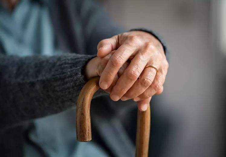 Elderly hand with wedding ring gripping wooden walking cane for support.