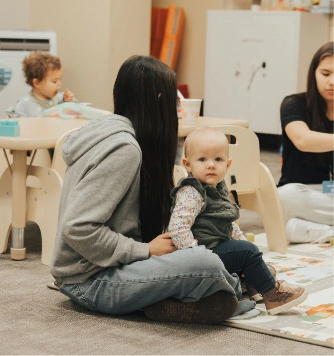 Parent sits with baby in cozy classroom with other children and teacher
