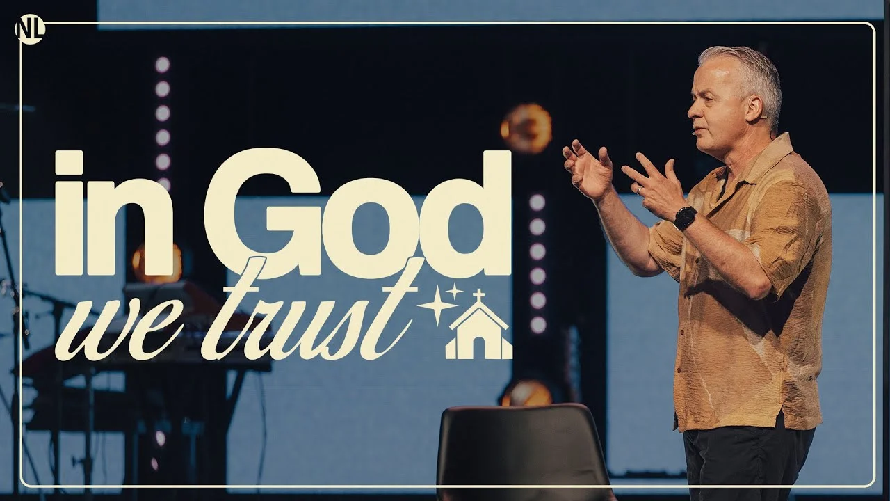 Man in tan shirt preaching with bold title "In God We Trust" beside church-themed icons on stage.