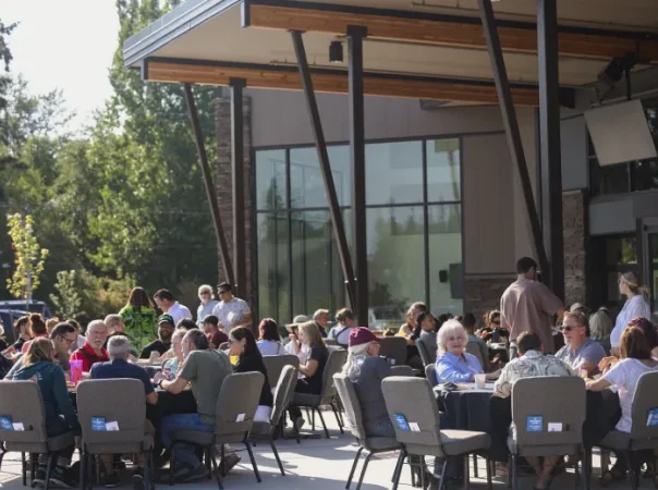 Group of people sitting together at outdoor tables under a shaded patio during a community event.