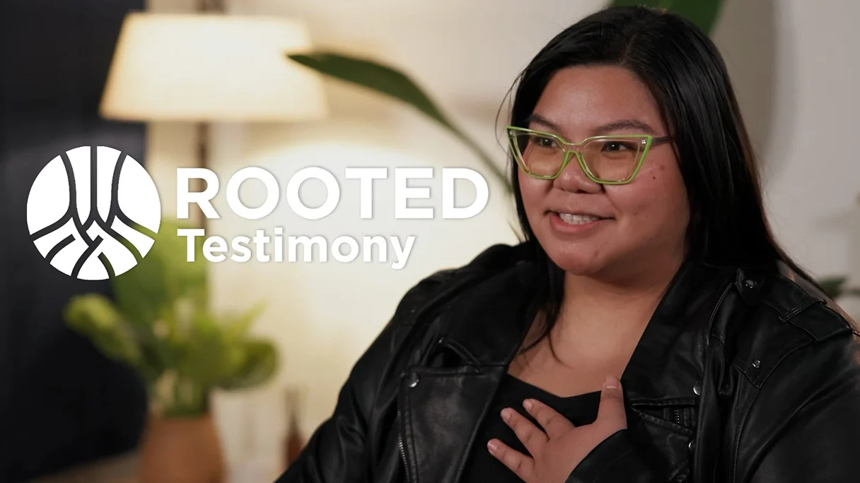Woman sharing testimony while seated indoors, wearing glasses and a black jacket, with Rooted Testimony logo displayed on the image.