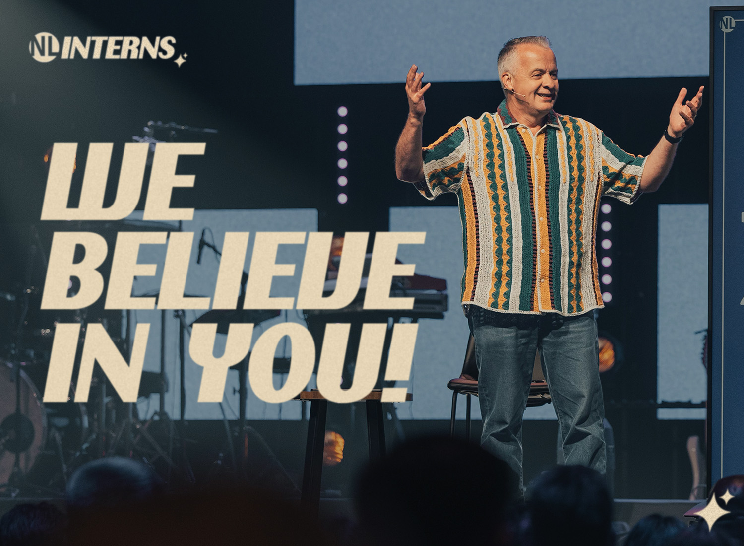 A smiling speaker in a colorful shirt gestures on stage beside bold text that says “We Believe in You!” during an NL Interns event.