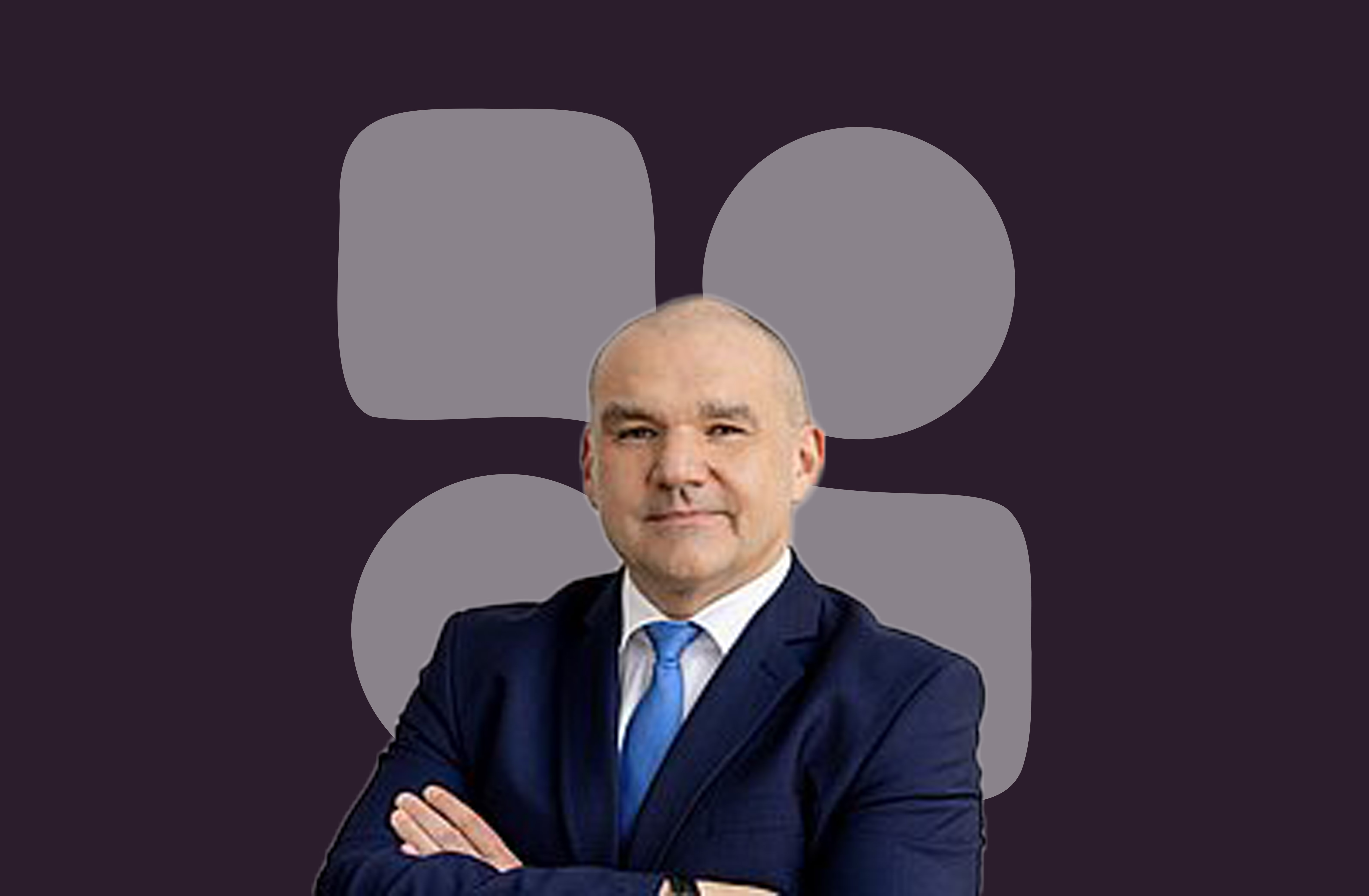 Bald man in a navy suit and blue tie with arms crossed, standing against a dark background with abstract rounded shapes.