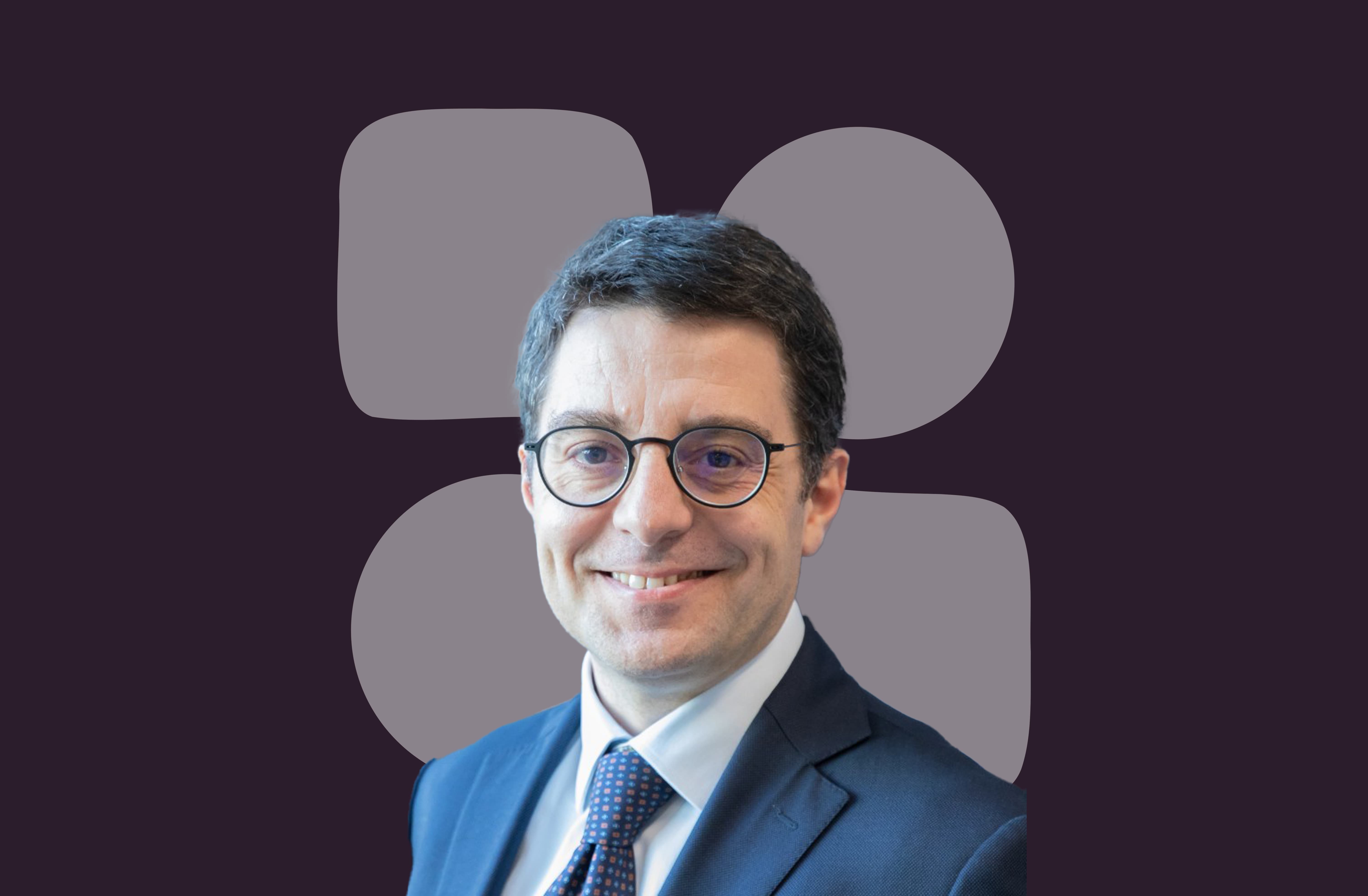 Man with short dark hair wearing glasses, a blue suit, white shirt, and patterned tie, smiling at the camera.