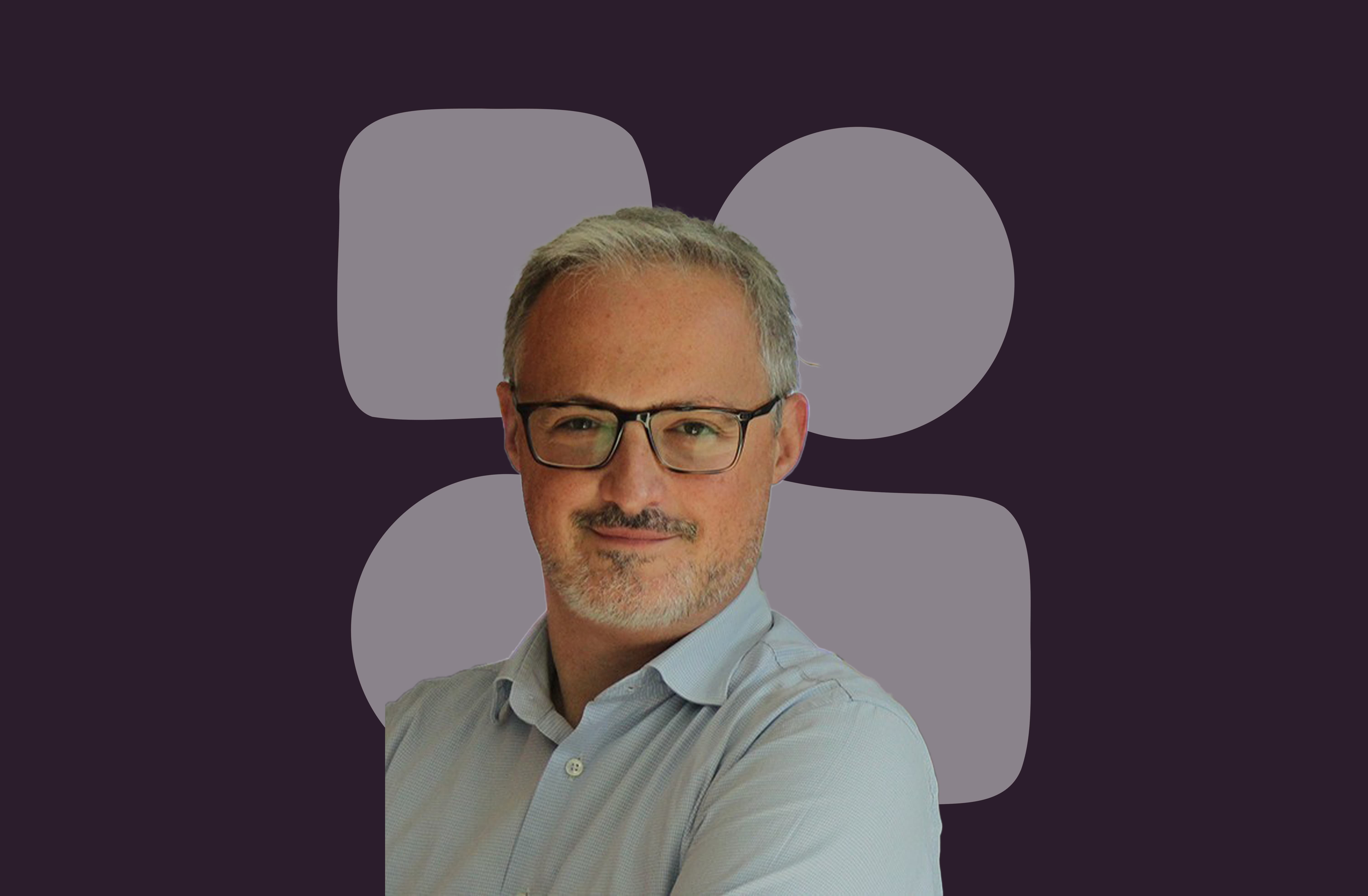 Man with gray hair and glasses wearing a light blue shirt, smiling with arms crossed against a dark background with abstract shapes.