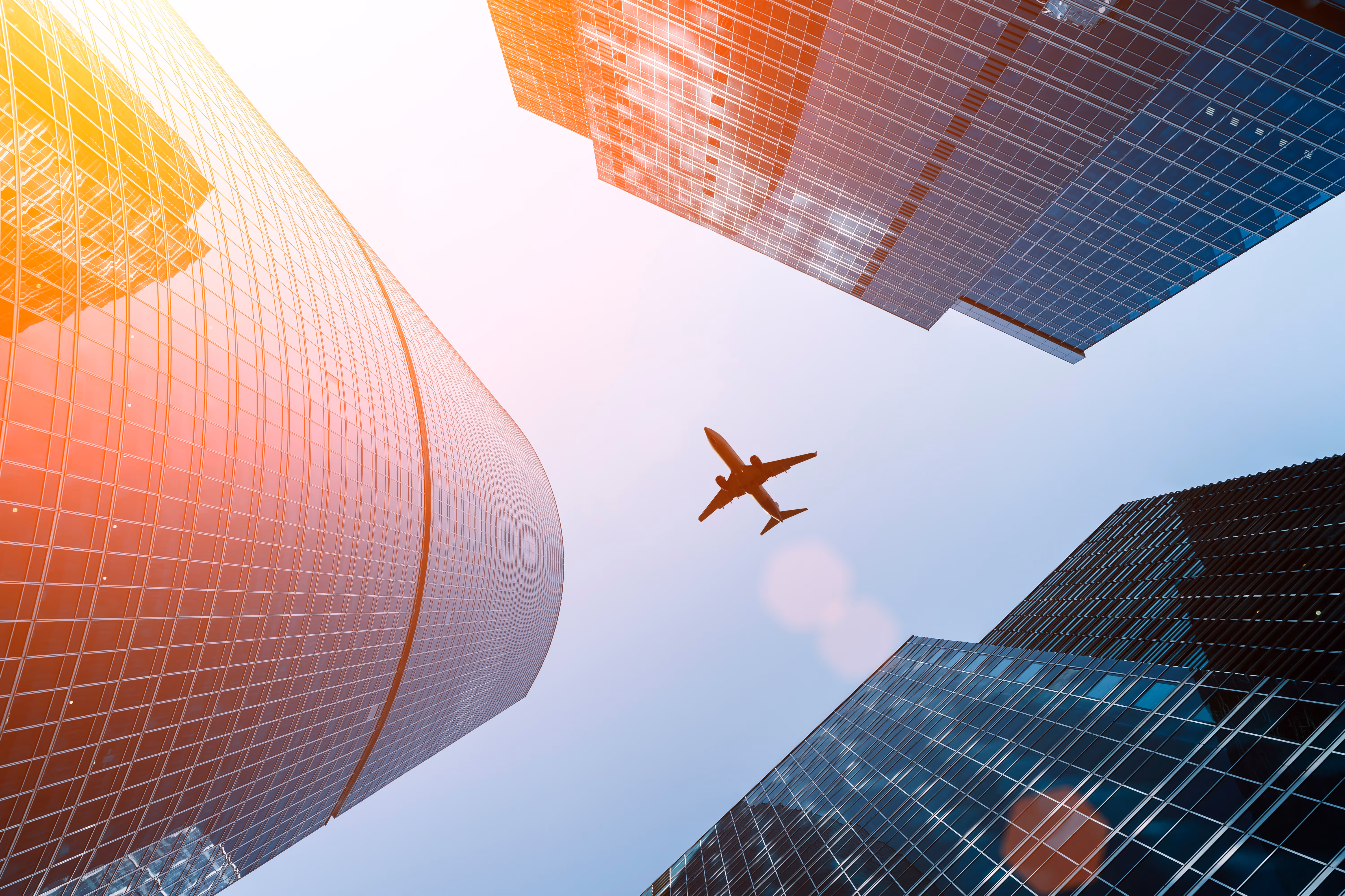 Airplane flying above tall glass skyscrapers with sunlight glare.