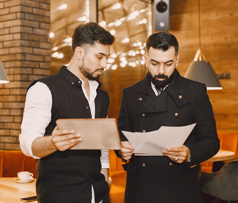 Two men in a modern restaurant or cafe discussing documents, one holding papers and the other a tablet.