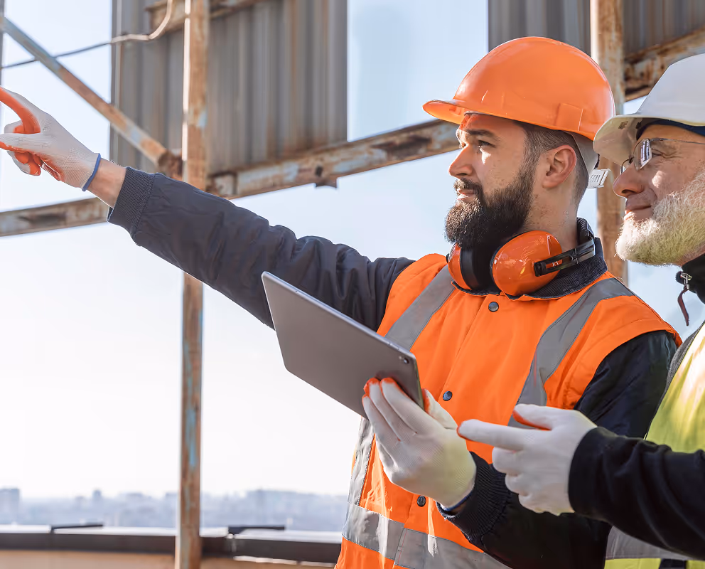 Two construction workers in safety gear pointing and reviewing plans on a tablet at a job site.