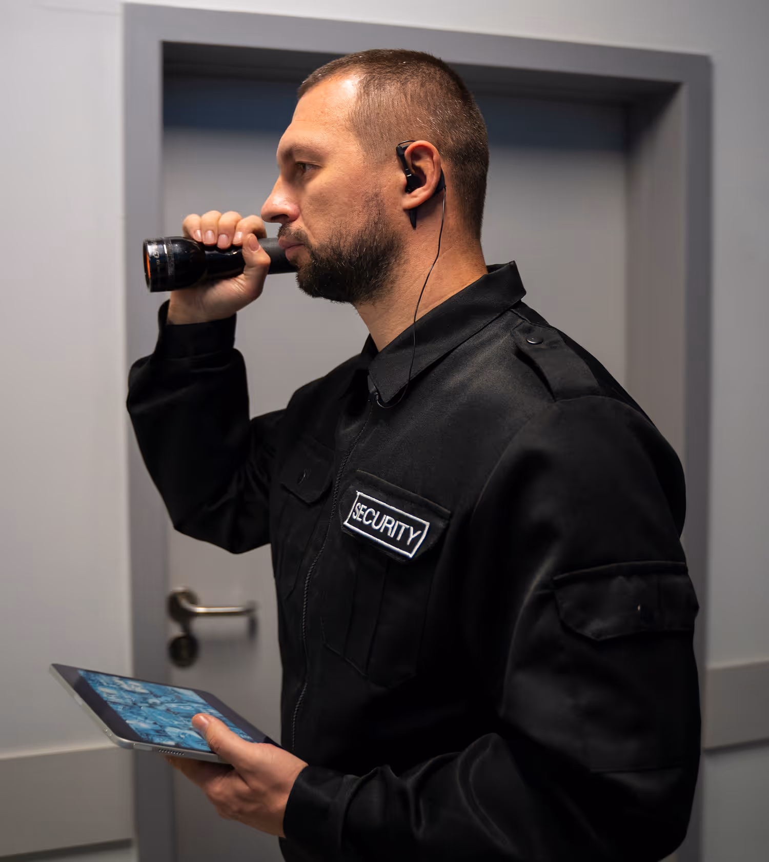 Security guard in a black uniform holding a flashlight to his eye and a tablet in his other hand while standing in a hallway.