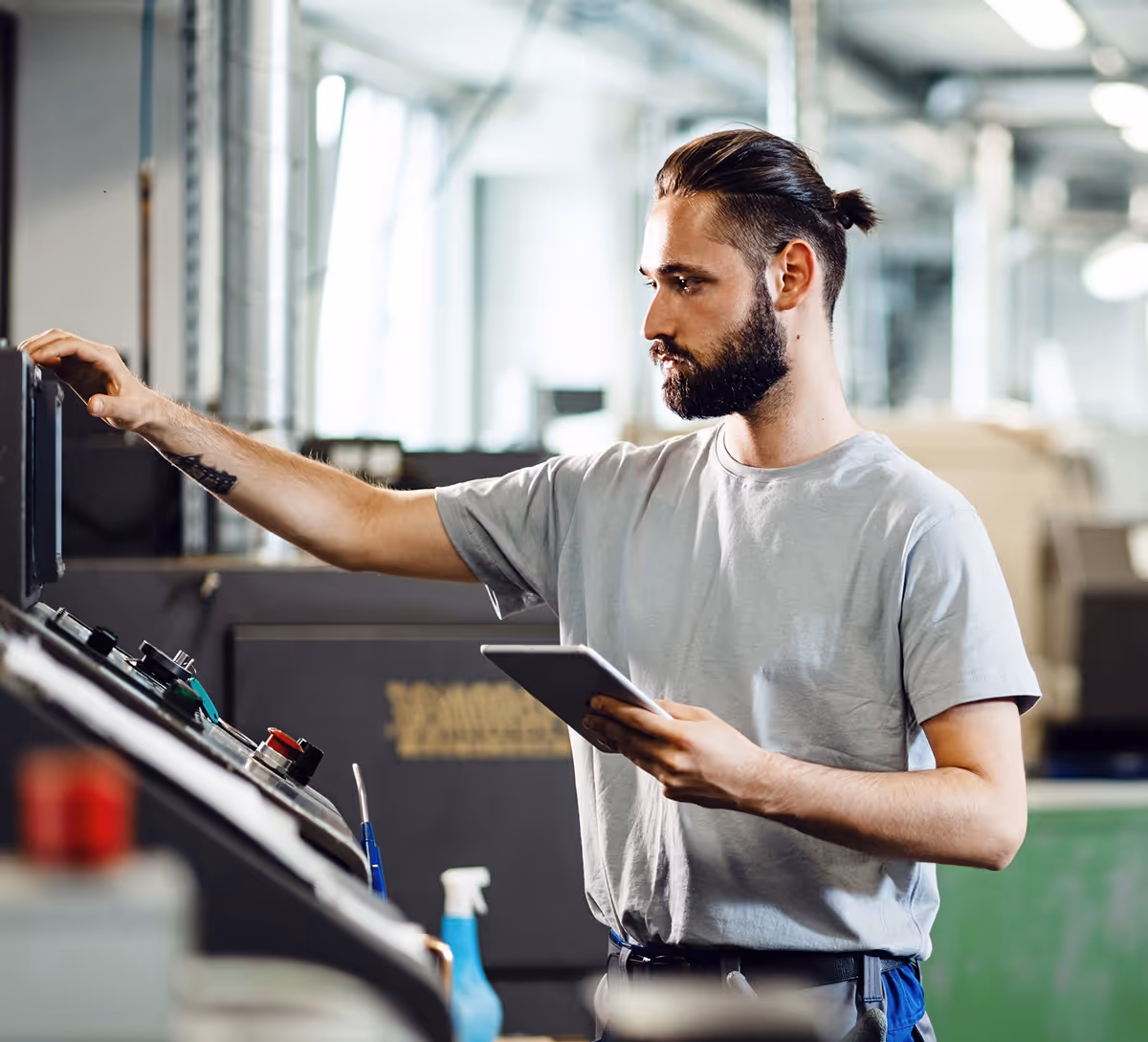 Industrial engineer operating a CNC machine while holding a tablet in a factory setting.