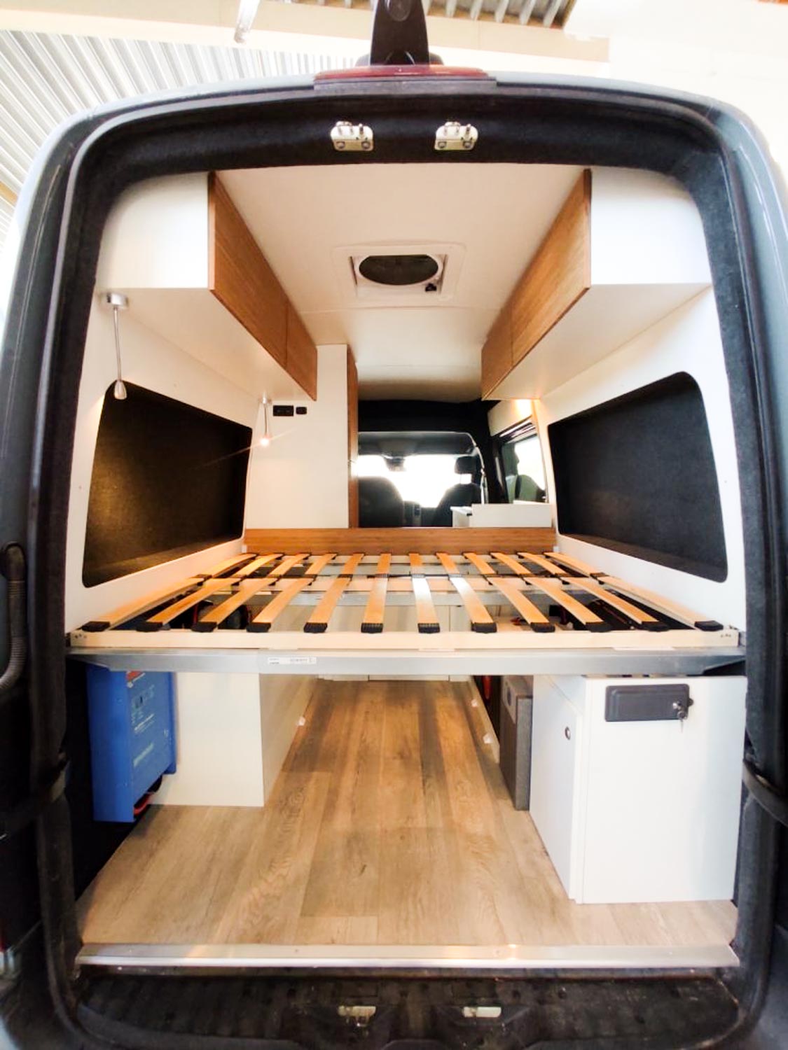 Interior view of a camper van showing a raised bed frame with wooden slats and storage cabinets on both sides.