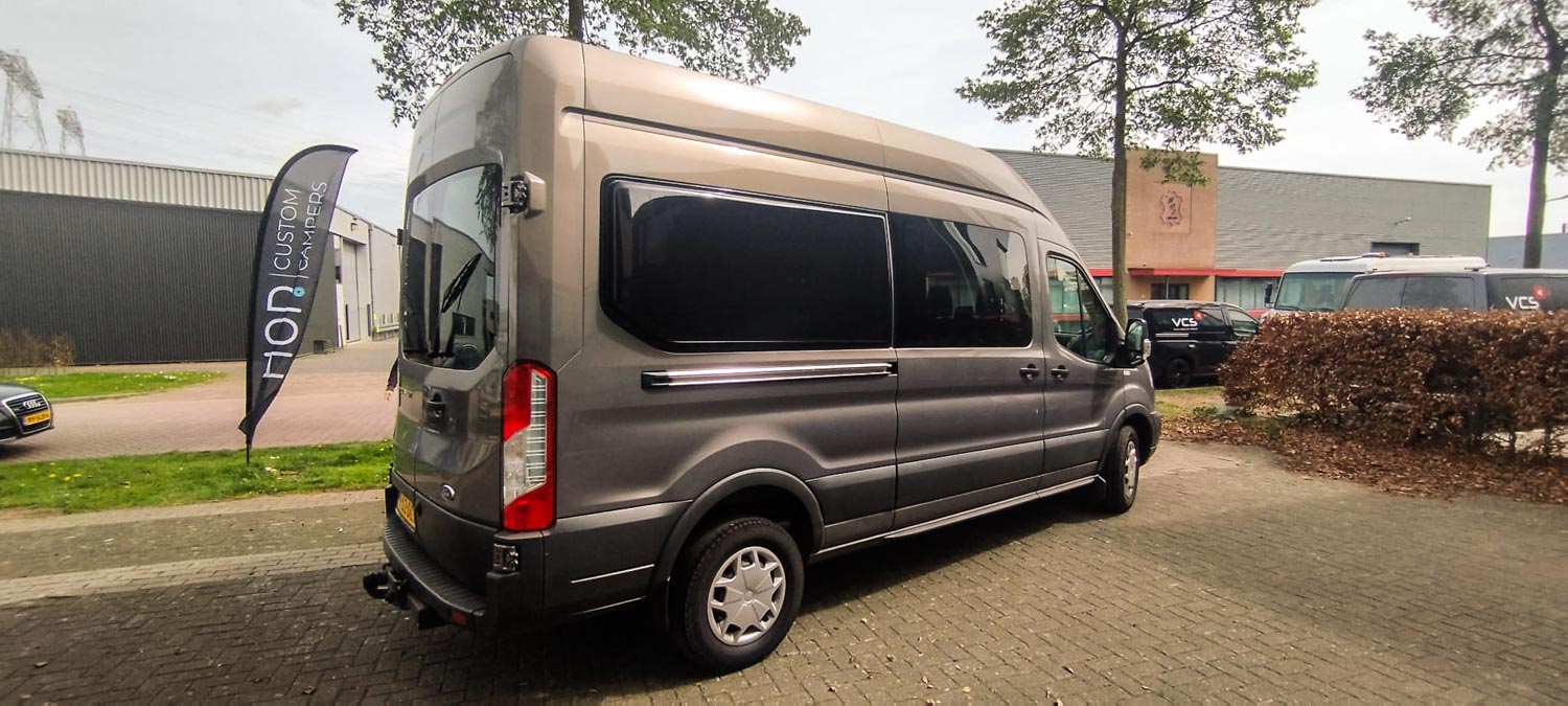 Gray high-roof camper van parked on a paved lot near a flag that says 'Custom Campers'.