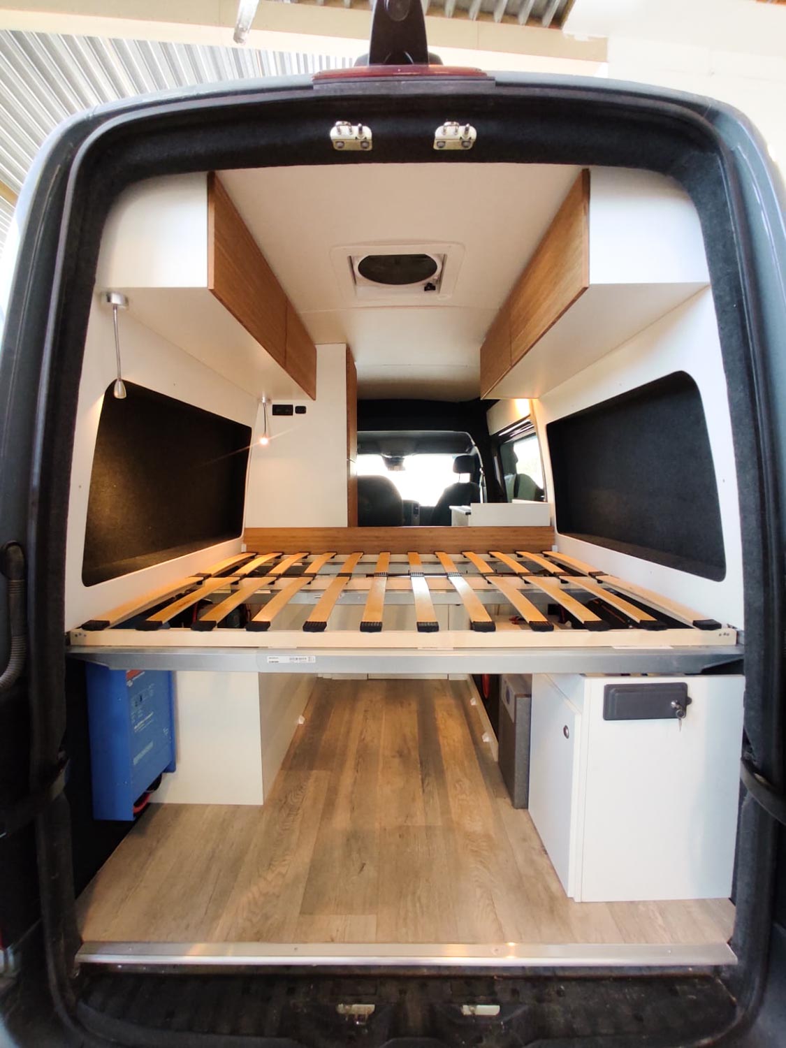 Interior of a camper van showing a wooden slatted bed frame, overhead cabinets, and light wood flooring.