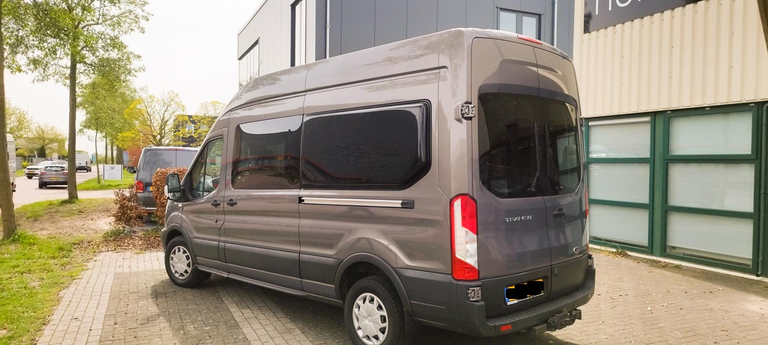 Gray Ford Transit van parked on a brick driveway beside a modern building and a tree-lined street.