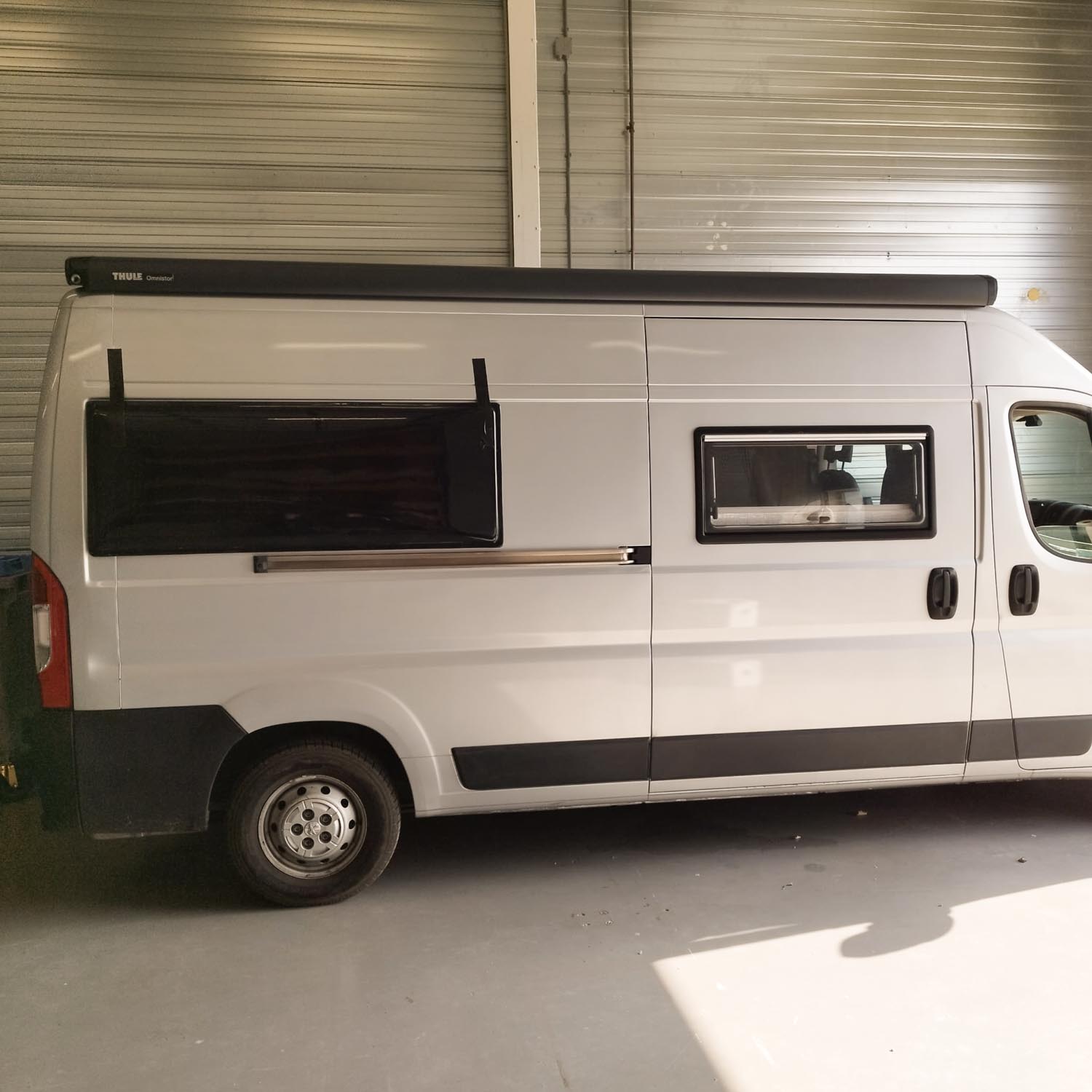 White camper van parked indoors with two black-framed windows and a Thule Omnistor awning mounted on the roof.