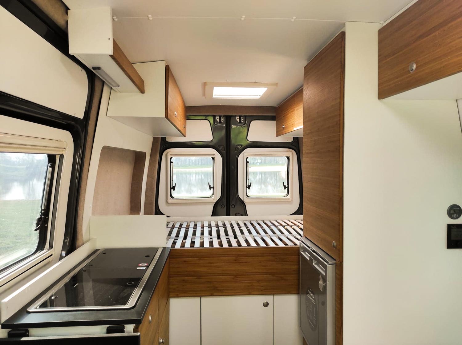 Interior view of a camper van showing a kitchen area with a stove on the left and a bed frame at the back with two rear windows.