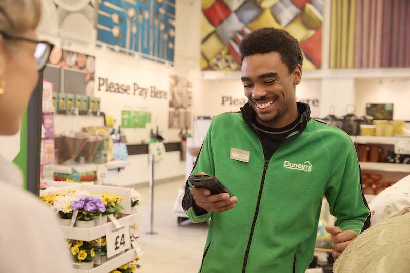 A Dunelm employee on the shop floor, smiling and a handheld computer. 