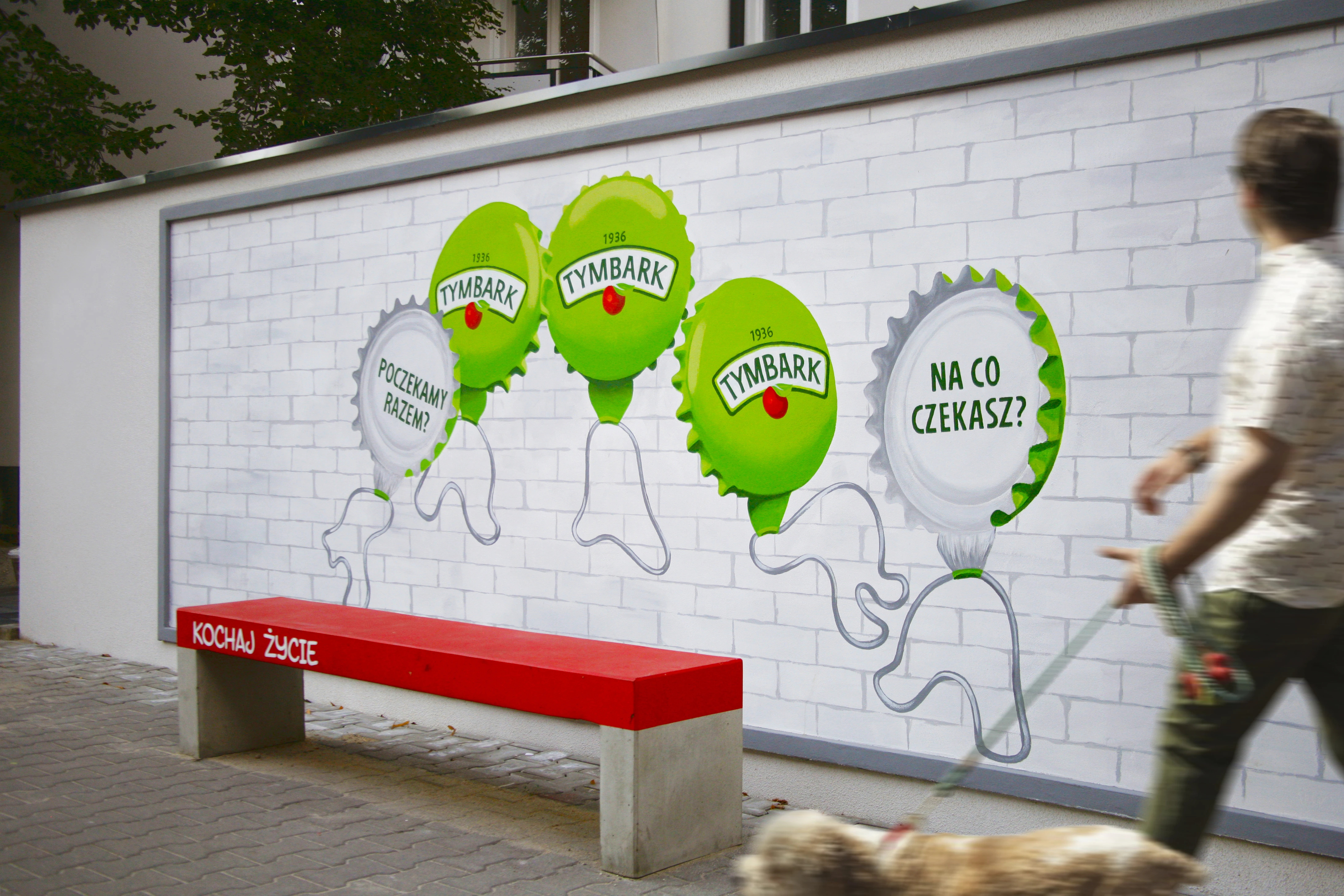 Mural Tymbarku namalowany przez SESME na ścianie w mieście. Obok człowiek idzie z psem