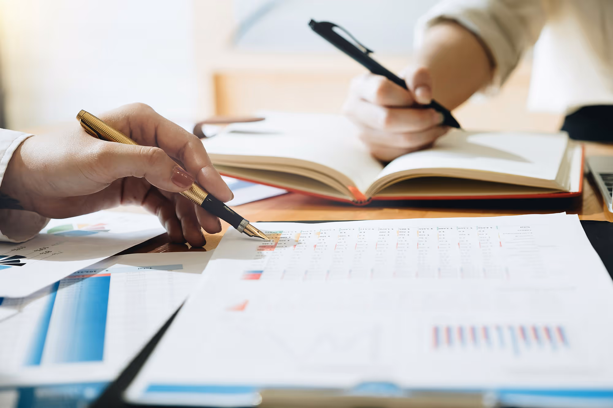 A hand holding a pen writing on a journal in the background and another hand with a pen on a paper in the foreground