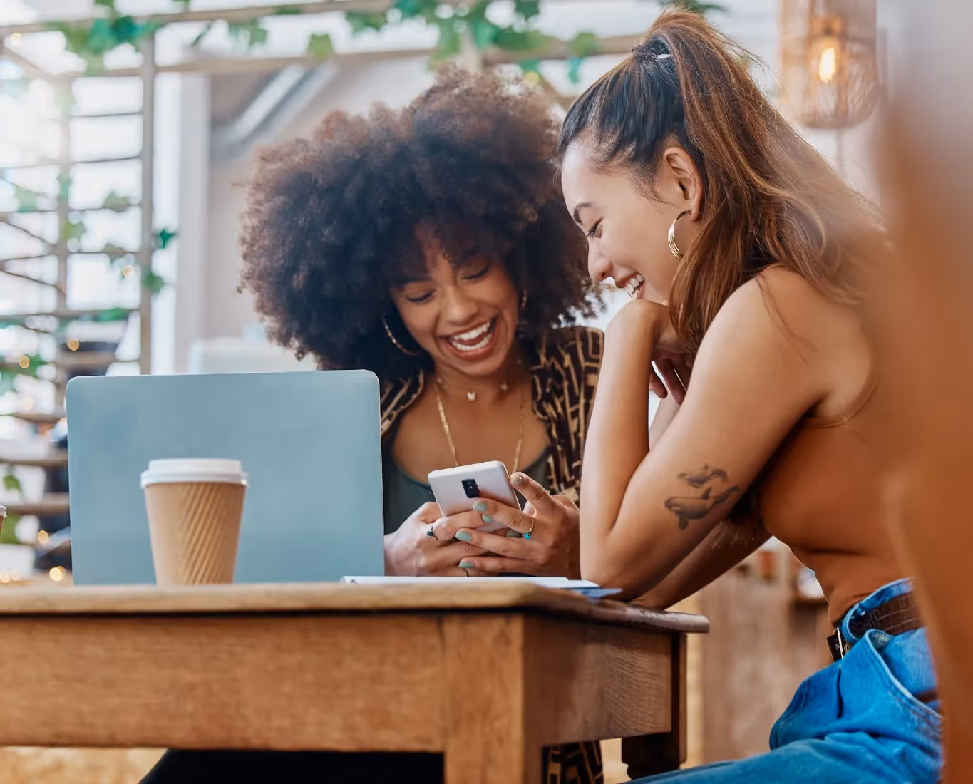 Two happy women laughing while looking at a smartphone and a laptop in a cafe, representing a convenient work-from-home and social space near Magnolia at Spring Hill in Spring Hill, Florida.