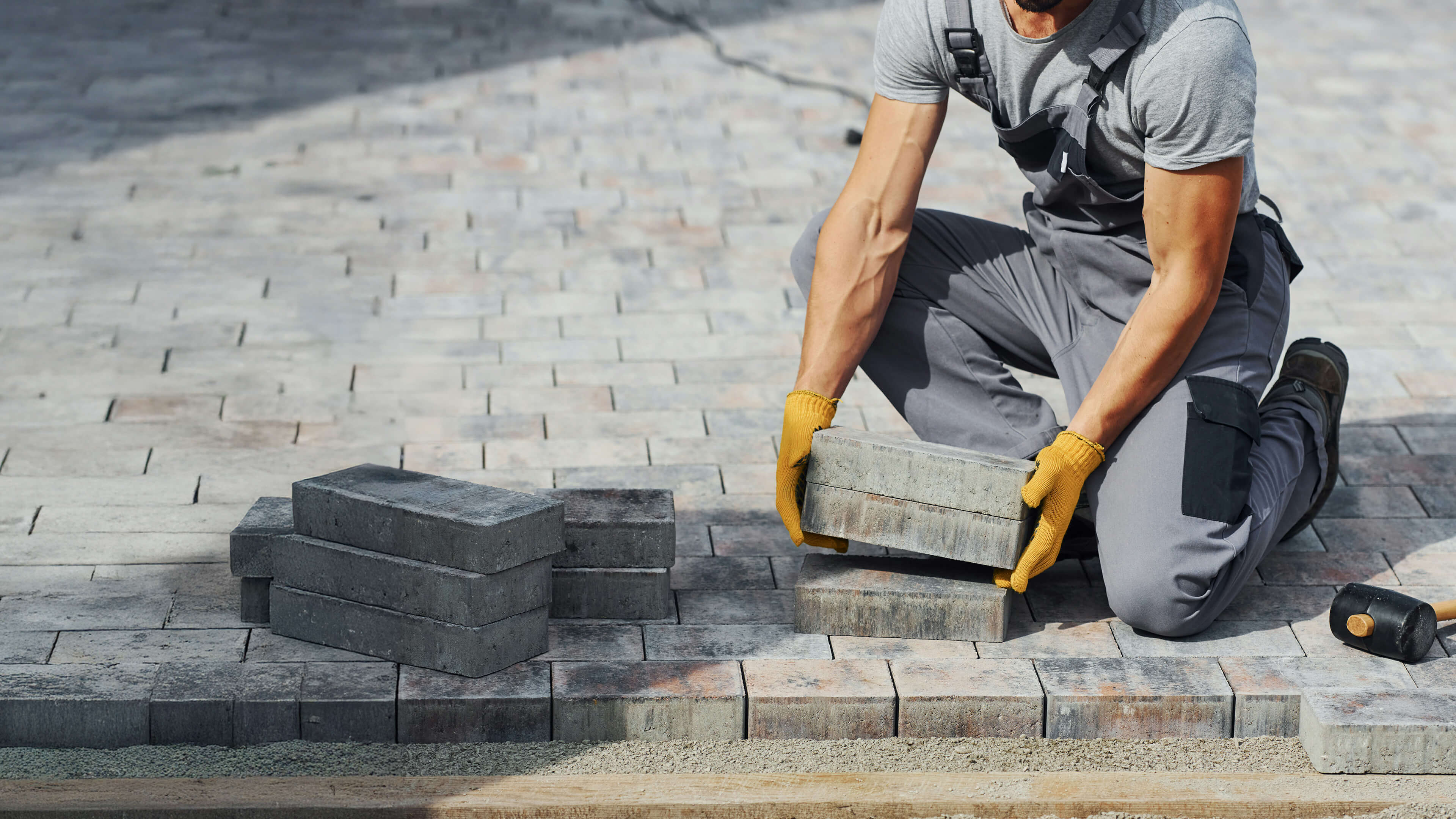 Worker in gray overalls and yellow gloves kneeling while laying concrete paving stones on a sand base.