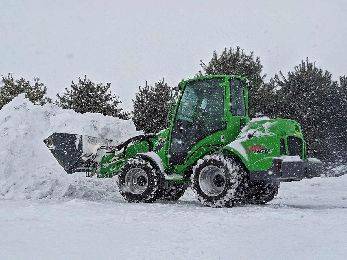 Green Avant 860 loader pushing a large pile of snow during snowfall with evergreen trees in the background.