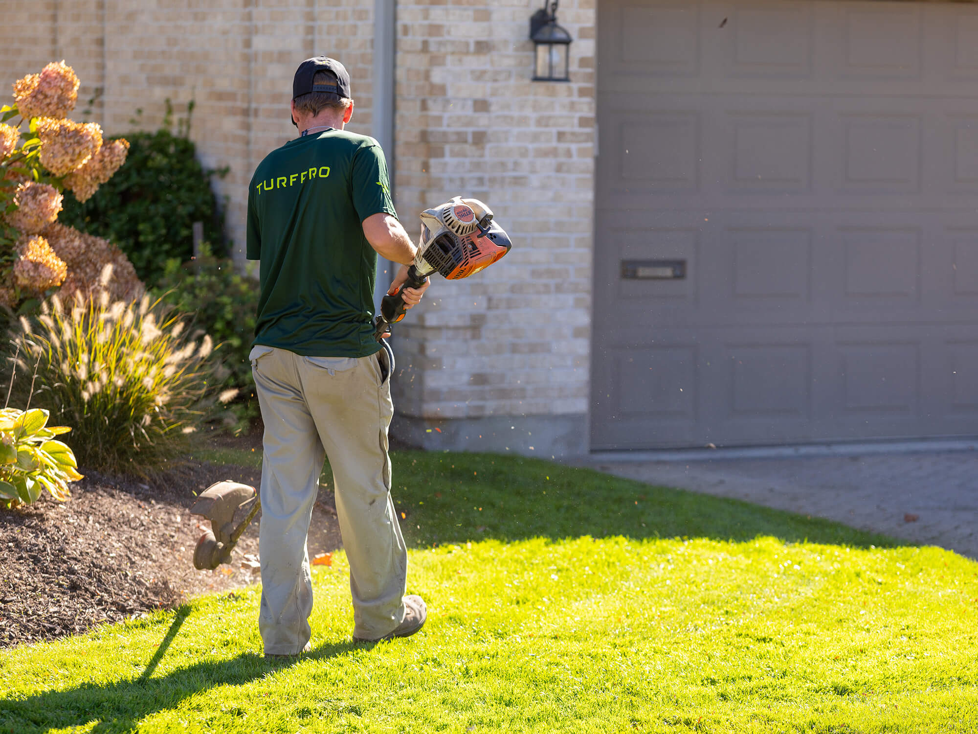 Landscaper wearing a green TURFFRO shirt using a string trimmer near a flower bed in front of a brick house.