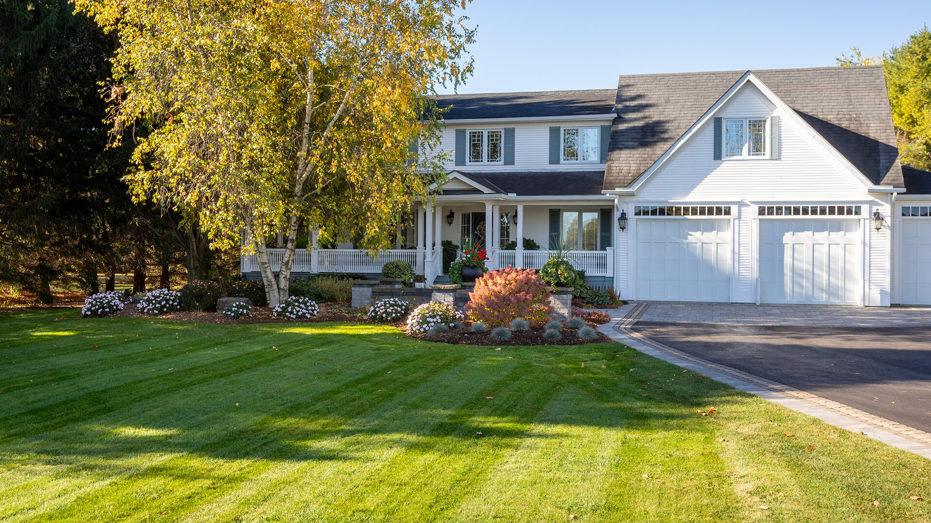 White two-story house with a large green lawn, landscaped garden, and a three-car garage on a sunny day.