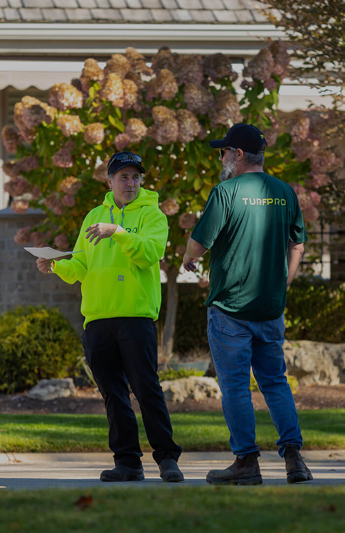Two men wearing TurfPro apparel stand outdoors on a residential street, one holding papers and gesturing while the other listens.