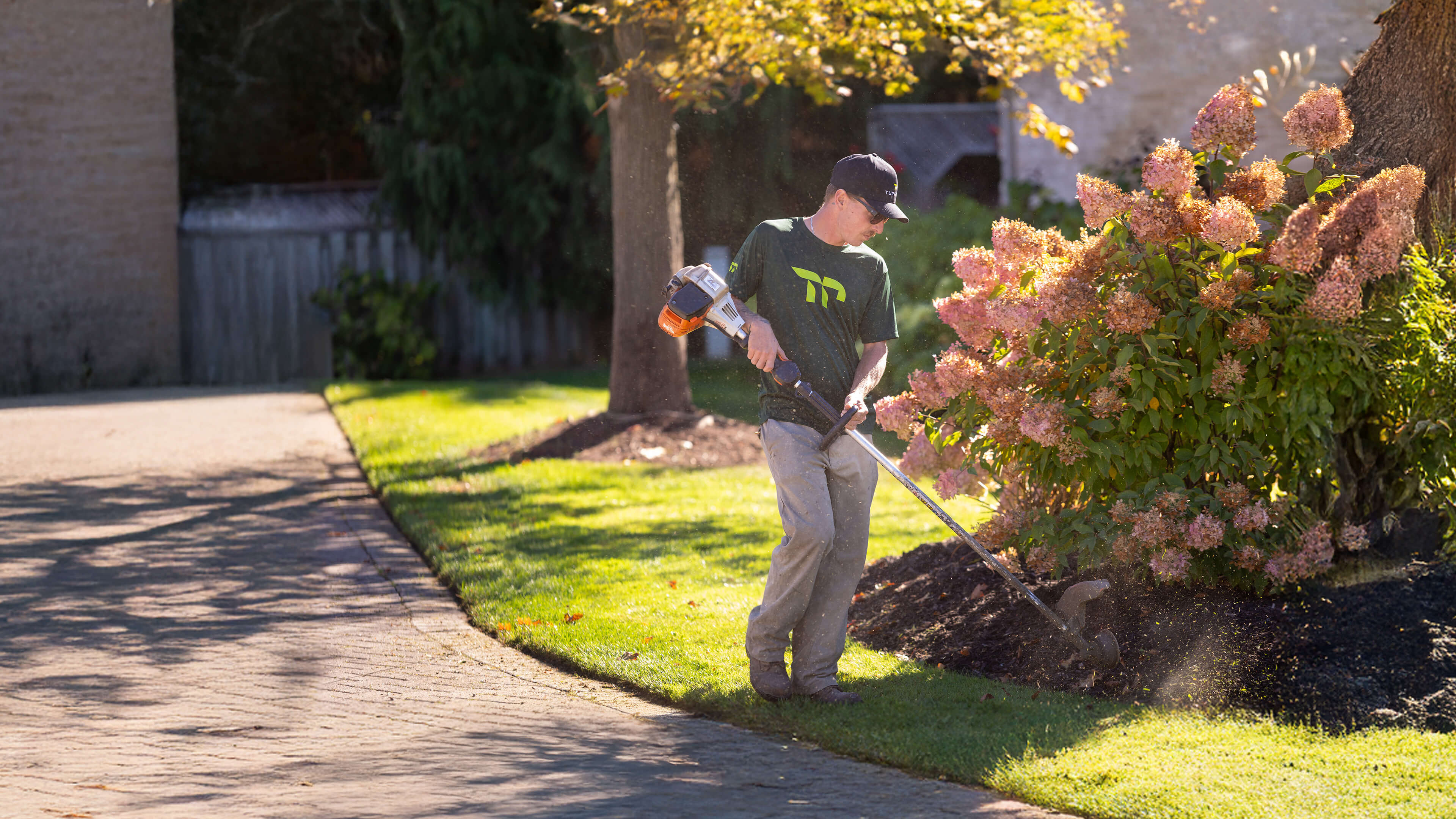 Man wearing sunglasses and a cap uses a string trimmer to edge grass near a flower bush beside a paved driveway.