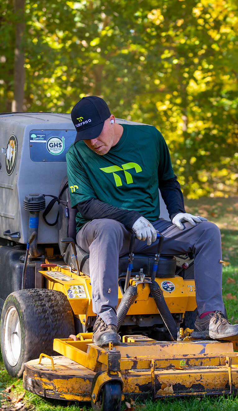 Man in a green Turfpro shirt and hat operating a yellow lawn mower on grass with trees in the background.