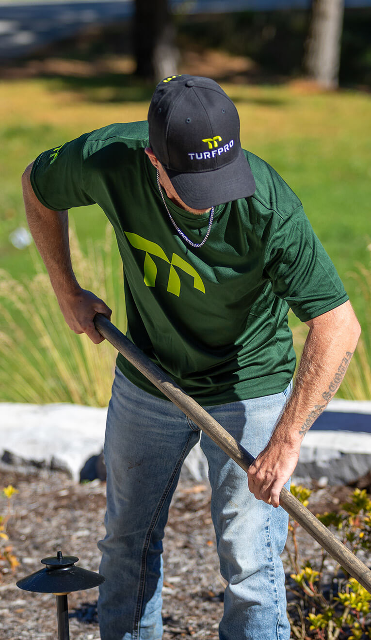 Person in a green TurfPro shirt and black TurfPro cap working outdoors with a wooden tool.