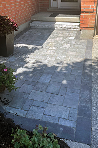 Stone patio with gray rectangular pavers bordered by darker bricks, leading to a small set of steps and a brick house entrance.