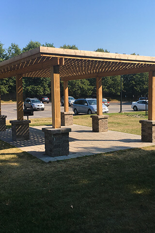 Wooden pergola with stone pillars located next to a parking lot under clear blue sky.