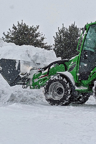 Green tractor with front loader carrying a large pile of snow during snowfall near trees.