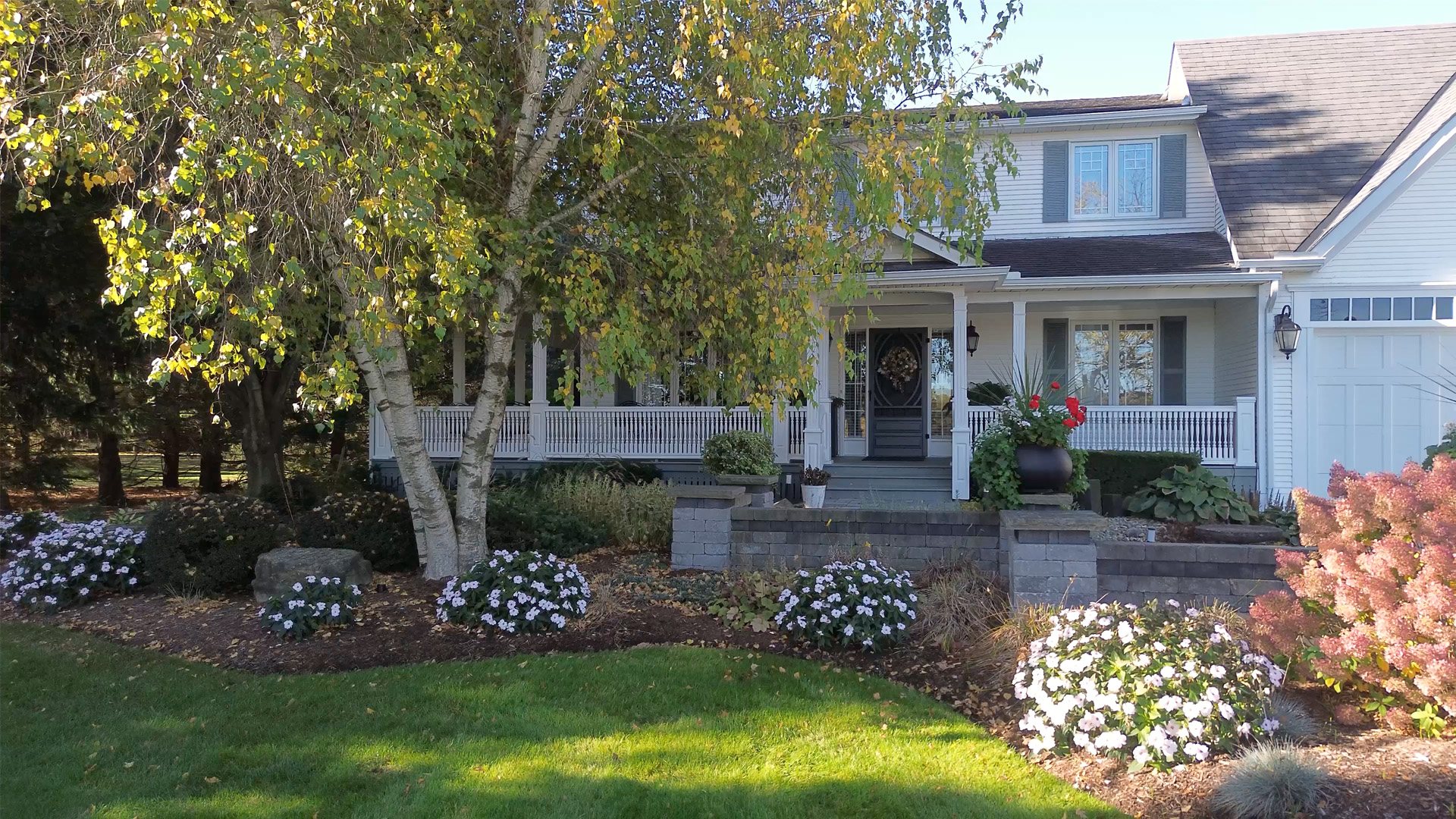 White suburban house with a front porch, surrounded by green lawn, flowering bushes, and a large tree with yellowing leaves.