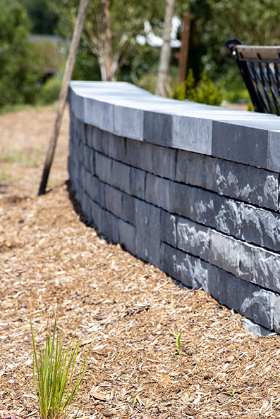 Brown textured retaining wall with gravel layer and a black metal railing on top, adjacent to a paved surface.
