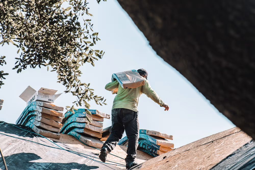 Roofer carrying a pack of asphalt tiles up a slanted roof.