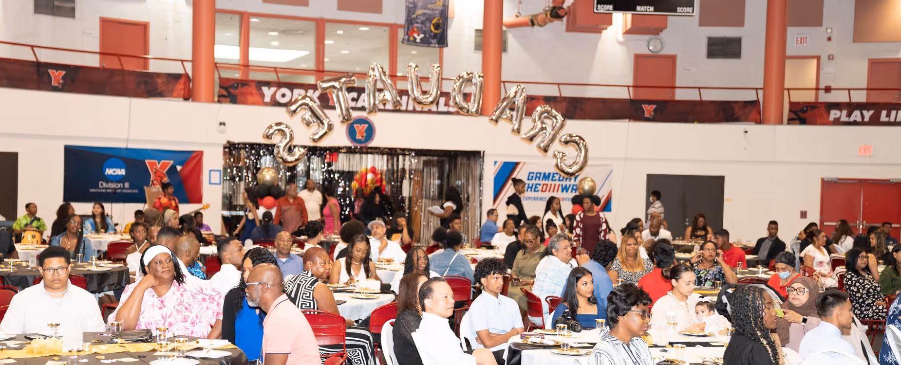 Community gathering in the school cafeteria—people eating and talking at decorated tables.
