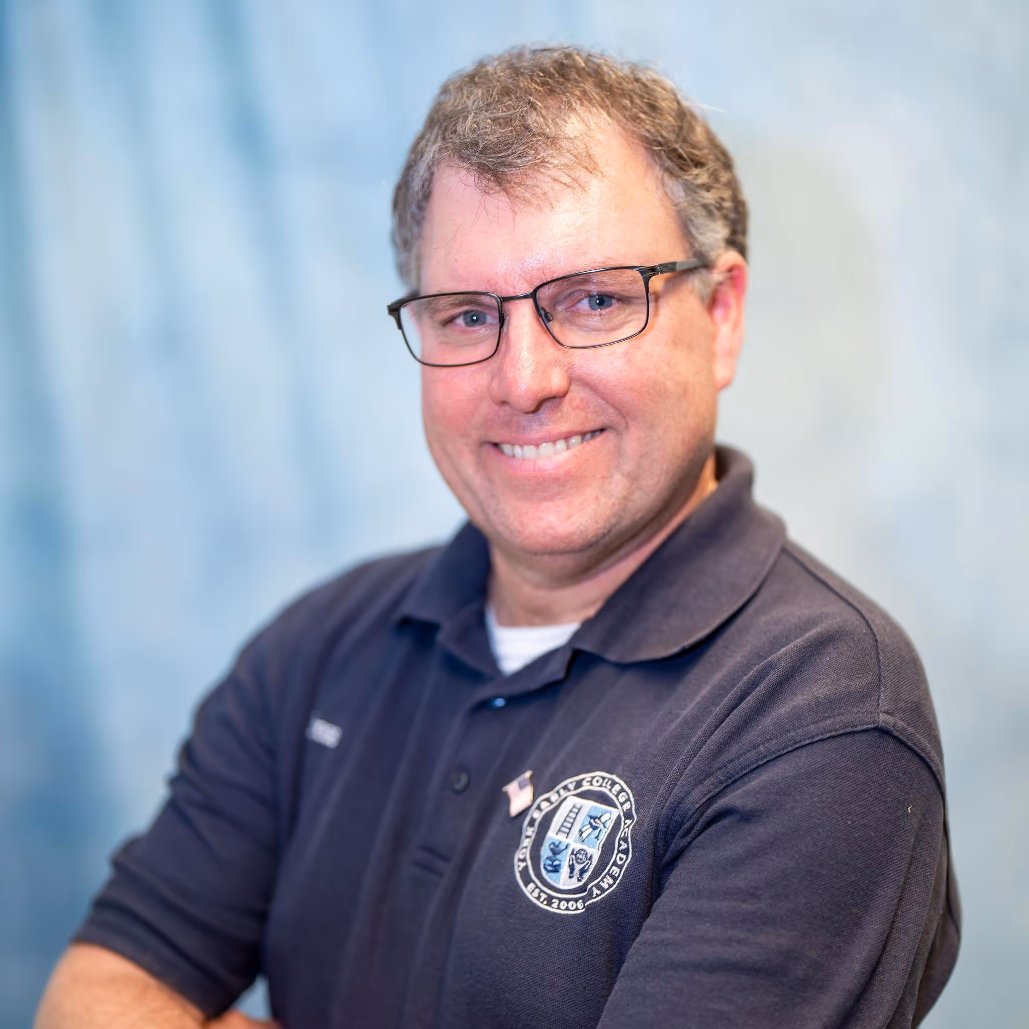 Portrait of a man wearing glasses and a dark polo shirt with an embroidered logo, smiling against a studio backdrop